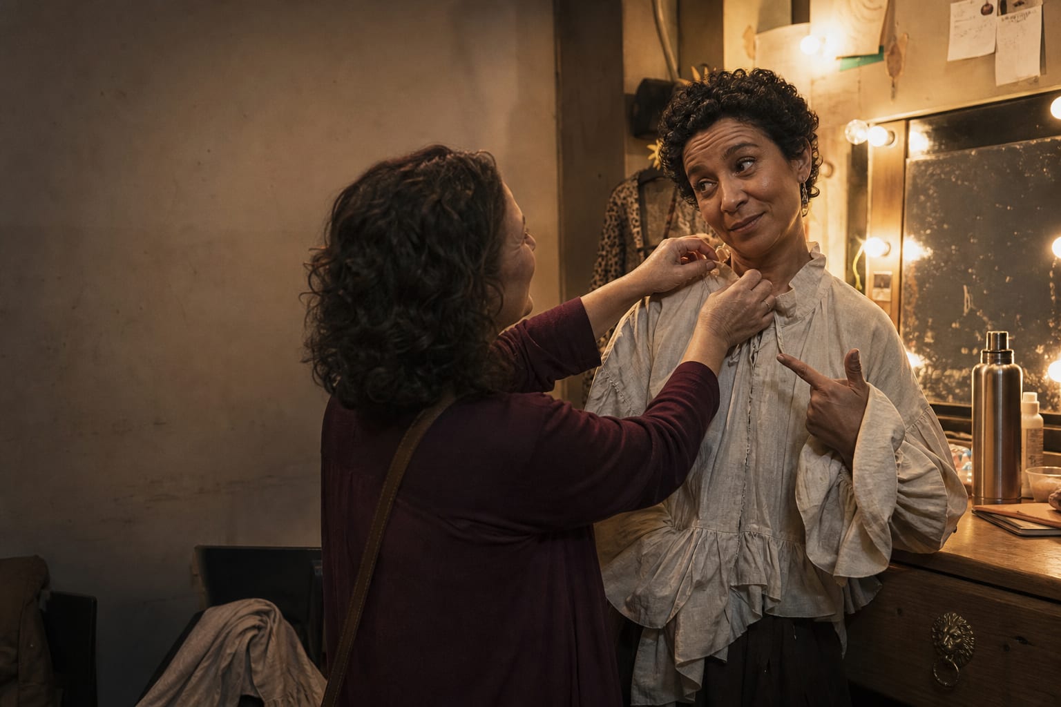 Duas pessoas se ajudam a preparar uma apresentação nos bastidores de um pequeno teatro